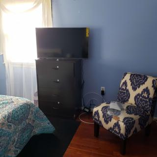 A cozy bedroom with blue walls features a television on a dresser, an armchair, and a bed near the window.