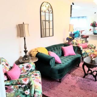 A cozy living room with a green velvet sofa, floral armchairs, and pink pillows, featuring a decorative mirror and lamp.