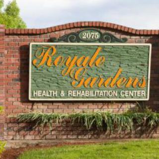 A sign for Royale Gardens Health & Rehabilitation Center, mounted on a brick wall with surrounding greenery.