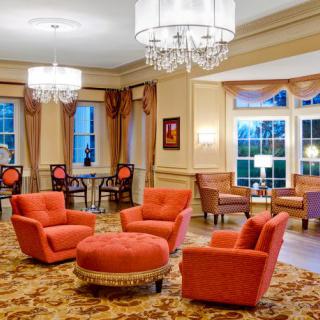 A luxurious lounge area featuring elegant chandeliers, plush armchairs, and a central ottoman in a warmly decorated room.