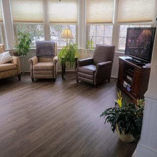 A cozy living room with comfortable seating and a TV, framed by large windows and houseplants.
