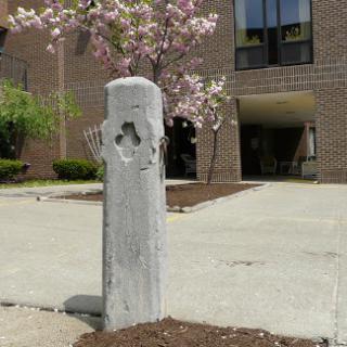 A weathered concrete post stands in front of a building, surrounded by blooming trees.