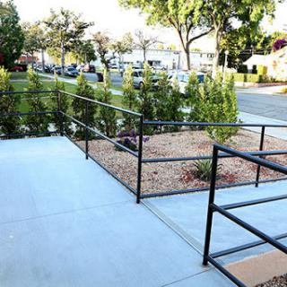 A view of a well-maintained outdoor area featuring a concrete walkway, black handrails, and neatly arranged greenery.