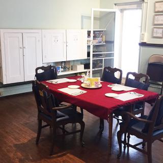 A dining room with a table covered in a red tablecloth, surrounded by chairs and set with placemats. The room features wooden floors and storage cabinets.