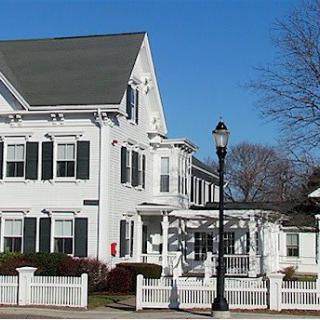 A charming white residential house with a gable roof and black shutters, surrounded by a white fence on a clear day.