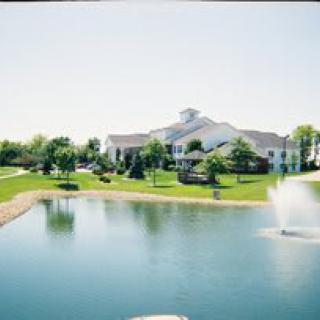 A scenic view of a large building near a pond with a fountain, surrounded by lush green landscaping and trees under a clear sky.