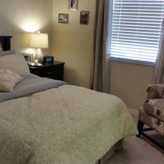 A cozy bedroom with a neatly made bed, bedside lamp, and floral chair by the window.