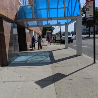 A sunny urban scene featuring a sidewalk with a blue-covered area and a bus stop.