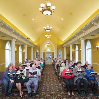 A group of people attending a church service, sitting in rows and holding hymnals inside a brightly lit chapel.
