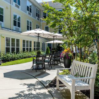 A serene outdoor patio area with tables, umbrellas, and benches surrounded by greenery.