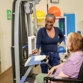 A healthcare professional assists a patient in a rehabilitation setting, demonstrating the use of exercise equipment.