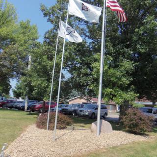 A display of flags in a landscaped area, surrounded by trees and a parking lot.