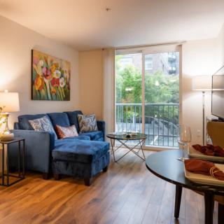 A cozy living room featuring a blue sofa, colorful artwork, and large windows allowing natural light.