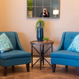 A cozy living space with two blue chairs and a small table featuring a vase, reflected in a wall mirror.