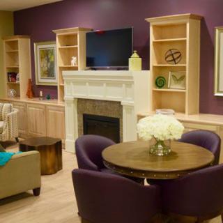 A cozy living room with a modern design featuring a fireplace, built-in shelves, and a television. The space is decorated with purple chairs around a round table adorned with white flowers.