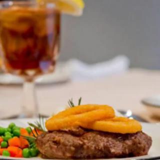 A delicious steak topped with onion rings, served with mixed vegetables and a glass of iced tea.