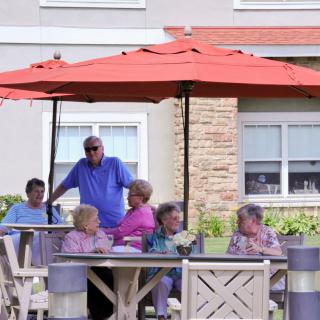 A group of seniors sitting around a table under a red umbrella, enjoying an outdoor conversation.