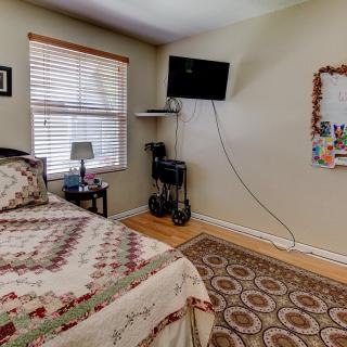 A cozy bedroom with a mounted television, patterned rug, and a neatly made bed next to a window.