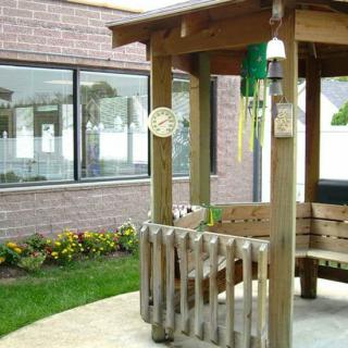 A small wooden gazebo with wind chimes is situated near a brick building, surrounded by a neatly maintained garden.