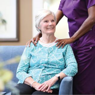 A caregiver provides support to an elderly woman sitting on a chair.