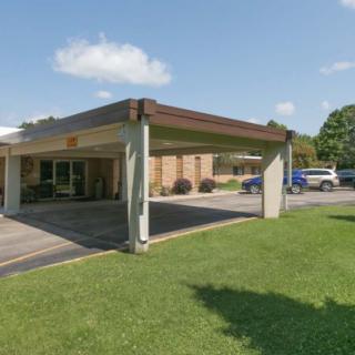 A building with a covered entrance and adjacent parking lot, surrounded by grass and trees under a partially cloudy sky.