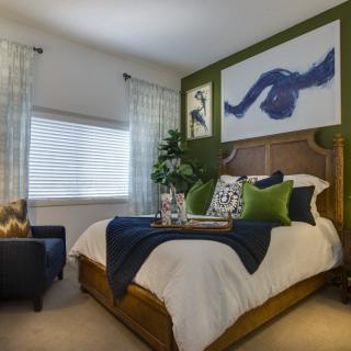 A stylishly decorated bedroom featuring a bed with green and blue accents, complemented by artwork on the walls and soft natural light filtering through the curtains.