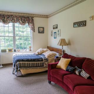 A cozy bedroom featuring a bed with patterned bedding, a red sofa with cushions, and a large window letting in natural light.