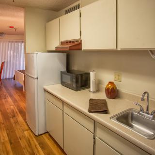 A compact kitchen in an apartment featuring cream-colored cabinets, a refrigerator, and a microwave on a wooden floor.
