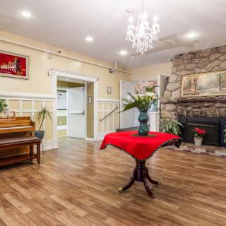 A cozy living room with a warm wooden floor, featuring a piano, stone fireplace, and a chandelier overhead. A small table with a red tablecloth showcases a decorative vase in the center.