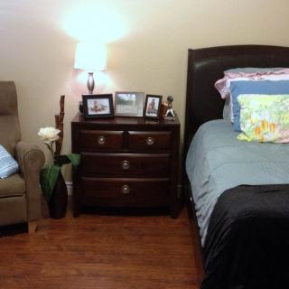 A cozy bedroom with a neatly made bed, nightstand with photo frames and a lamp, and an armchair with decorative pillows.
