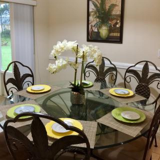 A dining area featuring a round glass table with ornate chairs, set with yellow and green themed place settings, and an orchid centerpiece.
