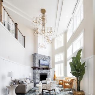 A spacious living room with high ceilings, featuring a modern chandelier and stone fireplace, complemented by large windows that allow natural light to illuminate the space.