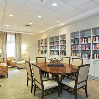A cozy library room featuring a round table surrounded by chairs, with bookshelves lining the walls and warm lighting creating an inviting atmosphere.