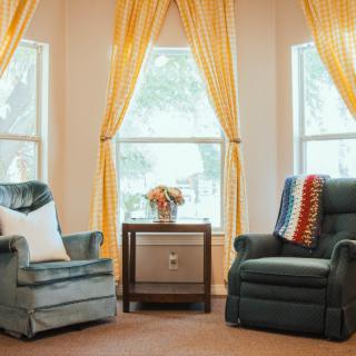 A cozy living room with two armchairs, a side table, and bright yellow curtains framing three large windows.