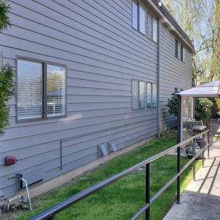 A side view of a house with gray siding, displaying windows and a grassy area bordered by a paved pathway and black railing. Outdoor furniture is placed under an awning.