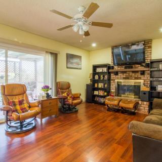 A cozy living room with wooden flooring, featuring a fireplace, television, armchairs, and a ceiling fan.
