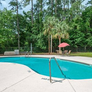 An outdoor swimming pool surrounded by lush greenery, with patio furniture and a sun umbrella nearby.