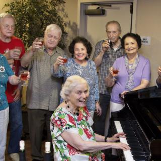 A group of elderly people celebrating with drinks while a woman plays the piano.