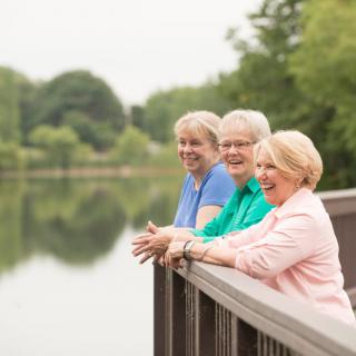 Three women enjoy a pleasant day outdoors, standing by a lakeside railing.