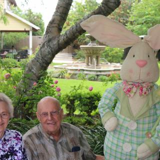 An elderly couple sits in a garden next to a large stuffed rabbit.