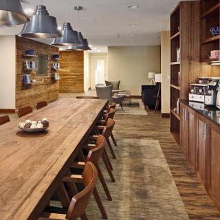 A modern dining area with a long wooden table, surrounded by chairs and illuminated by pendant lights, featuring a coffee machine and contemporary decor.