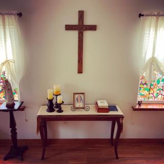A serene interior space featuring a wooden cross on the wall, surrounded by decorative elements such as candles and a table with personal items.