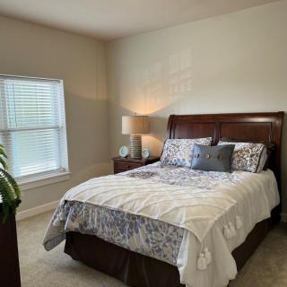 A cozy bedroom featuring a neatly made bed, bedside lamp, and window with blinds.