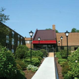 A building entrance with a red awning, surrounded by lush greenery and lamp posts under a clear blue sky.