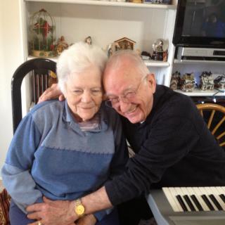 An elderly couple enjoying a warm embrace and smiling, seated next to a keyboard in a cozy home setting.