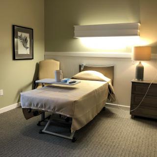 A well-lit hospital room featuring a neatly made bed, bedside table with a lamp, and framed art on the wall.