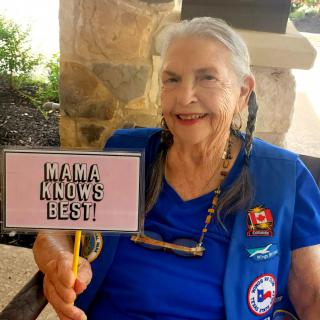 An elderly woman smiling while holding a sign that says 'Mama Knows Best.'
