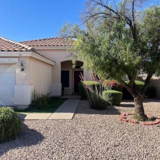 A single-story house with desert landscaping, featuring a tree and a pathway leading to the entrance under a clear blue sky.