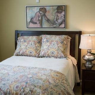 A cozy bedroom with a neatly made bed, decorative pillows and duvet, a bedside lamp, and a painting on the wall.