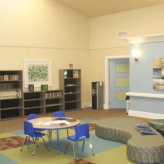 A well-organized classroom with small tables and blue chairs, surrounded by bookshelves and a play area.